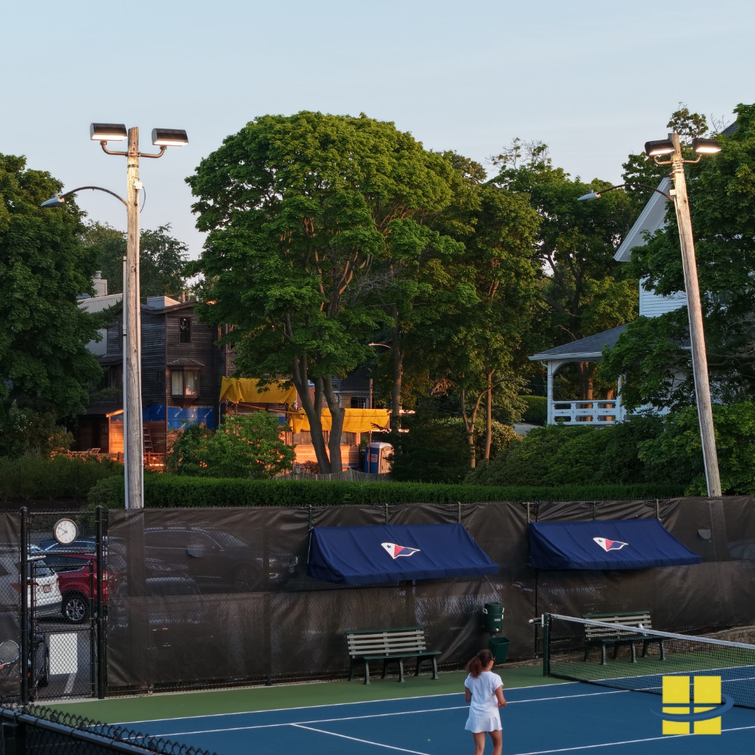 Ground-level dusk view of lit tennis courts at the Corinthian Yacht Club in Marblehead, MA — two players on court with APTI 600 LED sport lights on wood poles and neighboring residential properties visible behind the courts