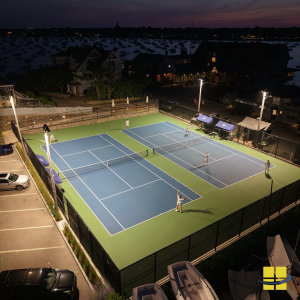 Elevated night view of twin LED tennis courts at the Corinthian Yacht Club in Marblehead, MA — six players in action under APTI 600 sport lights with Marblehead Harbor and moored boats visible in the background