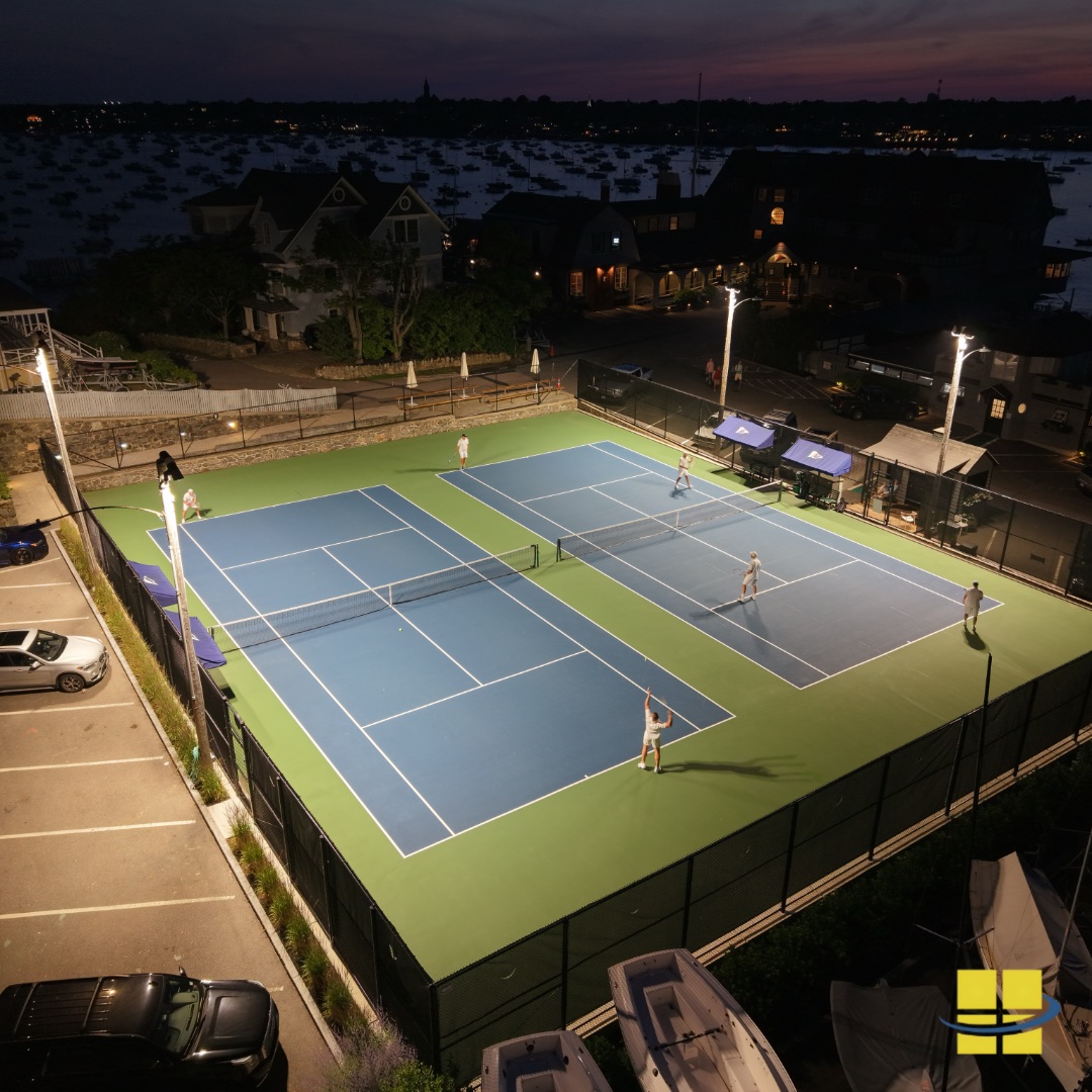 Elevated night view of twin LED tennis courts at the Corinthian Yacht Club in Marblehead, MA — six players in action under APTI 600 sport lights with Marblehead Harbor and moored boats visible in the background